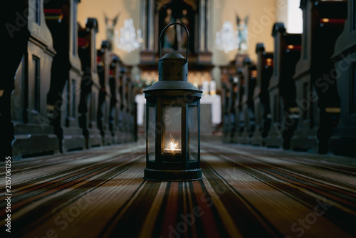 A lantern stands in the center of an empty church, casting a warm light. The wooden pews are lined up on both sides, creating a pathway. The setting shows a peaceful moment in time.