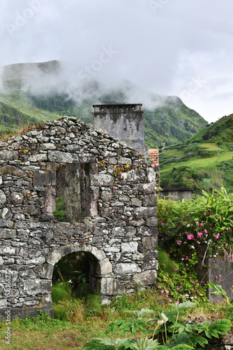 An ancient stone ruin of a village house overgrown with lush Azorean vegetation, set amidst a misty mountain landscape