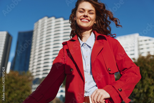 Fotografie Young woman in a red coat smiles broadly as she stands outdoors with modern city buildings behind, exuding confident, lively street style and a sunny, upbeat mood on a clear day