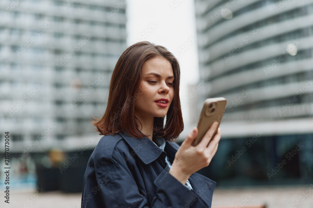 Fototapeta premium Young woman with straight brown hair wearing a dark coat looks at her smartphone with a slight smile in an urban environment with modern buildings in the background.