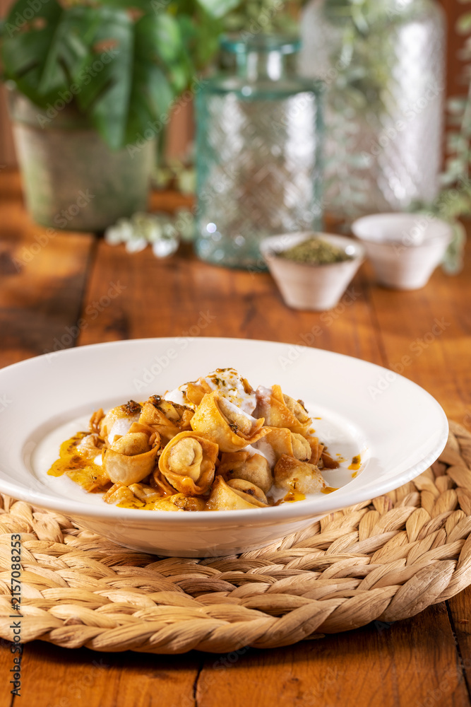 Fototapeta premium Traditional Turkish crispy fried manti (dumplings) topped with garlic yogurt and spiced chili butter sauce, served in a white bowl on a woven placemat over a warm wooden table.