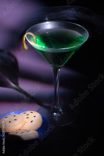 Festive halloween party pairing showing a green sparkling potion cocktail in a martini glass alongside a plate of bat sugar cookies and eerie smoke clouds on a dark purple table