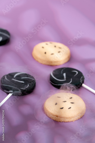 Halloween dessert table featuring bat cookies and black lollipops surrounded by mystical white smoke clouds on purple background, with copy space designed for spooky candy promos