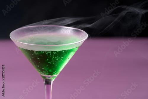 Close up of a sparkling green Halloween martini cocktail with a trail of white smoke vapor flowing across a purple background, creating a moody and design with space for text
