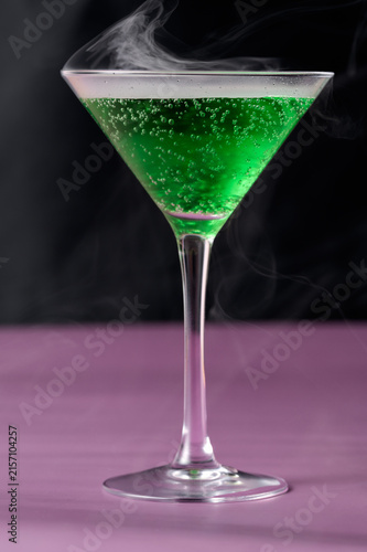 Vibrant neon green halloween potion cocktail in a martini glass garnished with a lemon twist, featuring thick white smoke mist rising from the glass over a vivid purple background