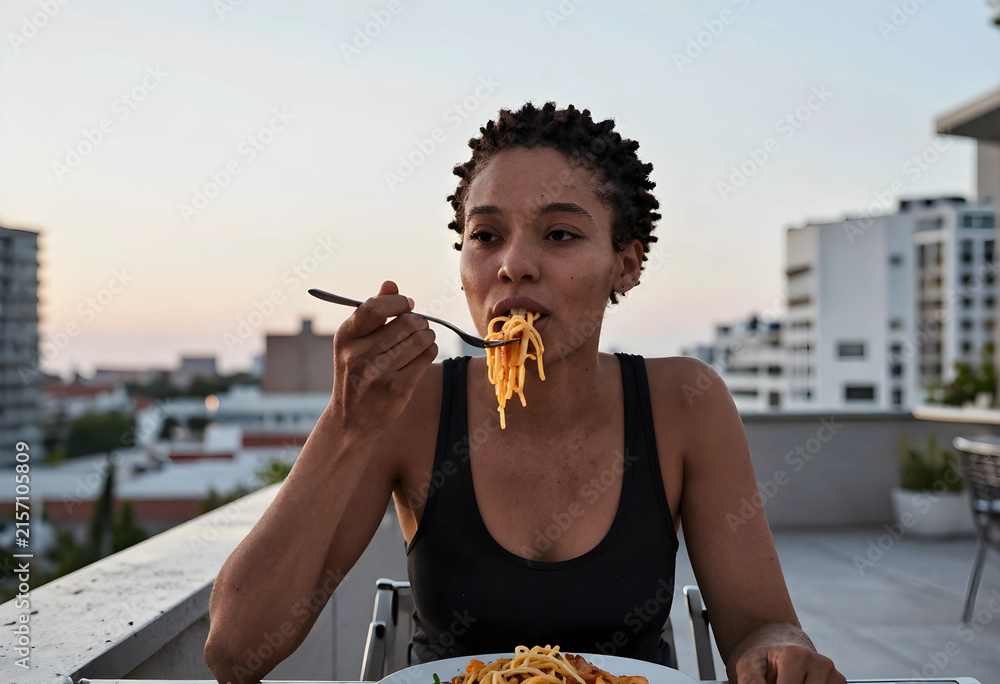 Fototapeta premium Young african american woman eating spaghetti with a fork on a rooftop terrace with city buildings in the background, the city buildings are visible in the background, creating an urban outdoor
