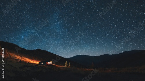 Small illuminated cabin beneath vast starry night sky over mountain
