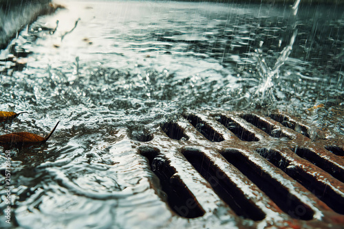 Rainwater flows into a storm drain hatch during a heavy downpour in a quiet urban area