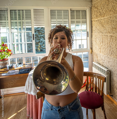 Young woman plays trumpet indoors beside large bright windows, framed by warm daylight, textured wall, wooden chair, and casual home setting, showing focused practice and musical performance