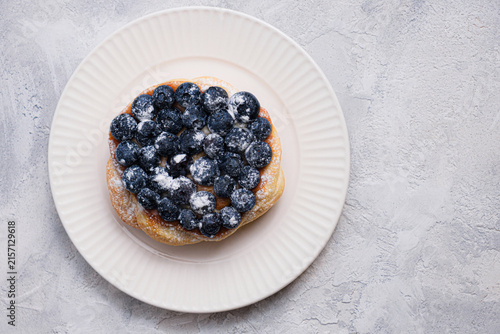 Top view of golden puff pastry Danish topped with fresh blueberries and powdered sugar on white ribbed plate, textured light gray background, copy space