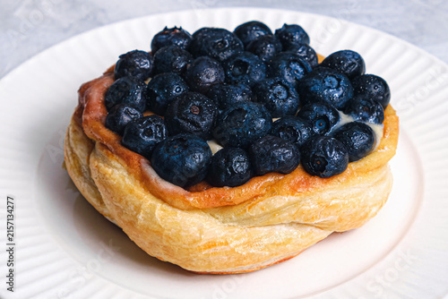 Close-up angle of golden puff pastry Danish piled with juicy fresh blueberries and cream filling on white ribbed plate, light textured background
