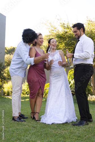 Diverse wedding group toasting on lawn, bride in bridal gown holding flute, man pouring champagne
