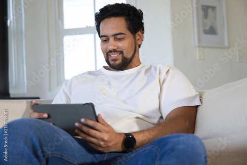 Mid adult African man wearing cream T-shirt sitting on beige couch in living room holding tablet