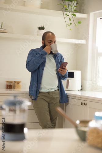African American man leaning in bright kitchen wearing denim shirt holding grey mug checking phone