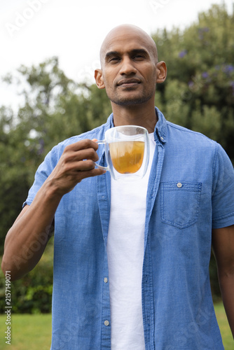 Adult African American man holding double-walled glass mug of tea standing in yard in denim shirt