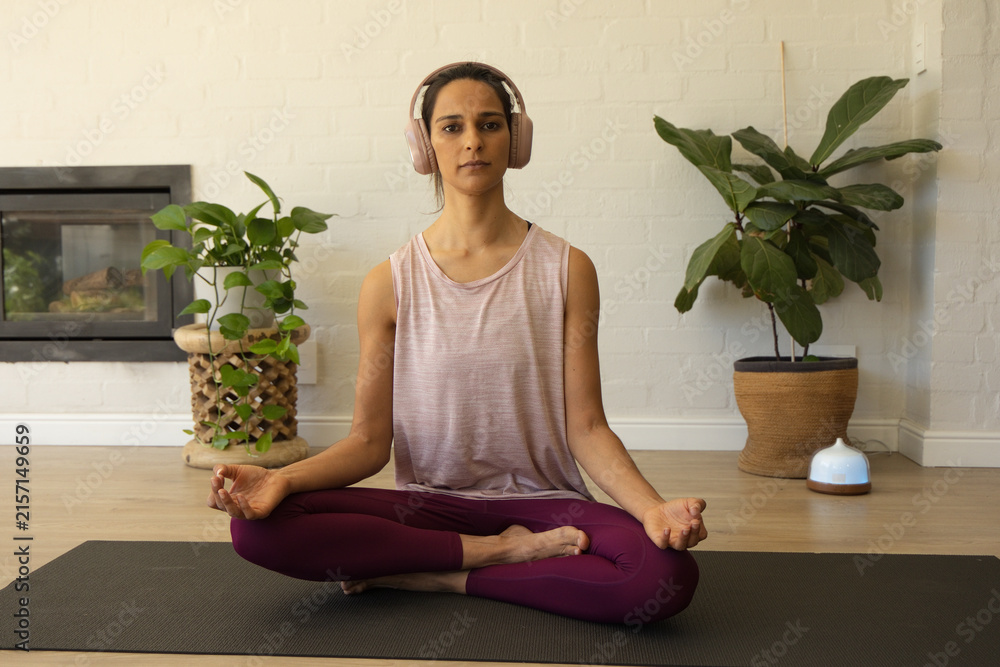 Fototapeta premium Female meditating on yoga mat at home wearing headphones, pink top, plum leggings with plants
