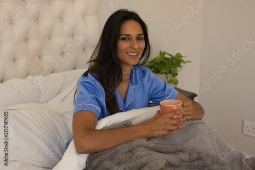 Indian woman sitting up on bed wearing light blue satin pajamas holding peach ceramic mug