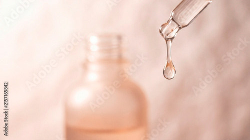 Macro photography of a glass dropper with a dripping liquid serum drop and a blurred frosted cosmetic bottle in a warm pink background for beauty skincare mockup.