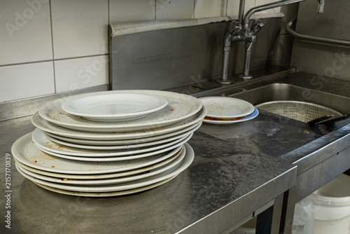 Stacked dirty white plates on counter at commercial kitchen sink area requiring dishwashing