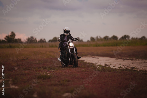 Motorcyclist in white helmet on motorbike with glowing headlight on countryside road at dusk. Night travel, safety and adventure lifestyle concept