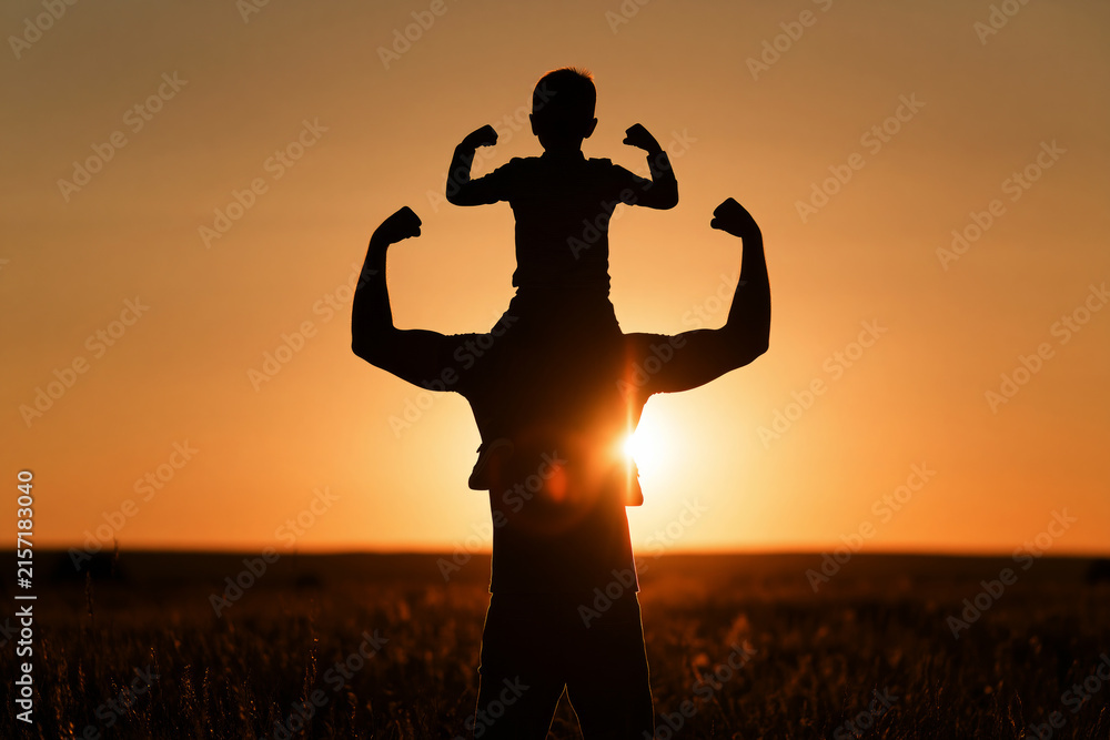 Fototapeta premium dark silhouette of a father carrying his young son on his shoulders in an open grassy field at sunset,