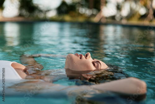 Pool scene with a woman floating peacefully, relaxation evident as she leans back, water shimmering, calm reflections on the surface, a serene lifestyle moment by the outdoor pool