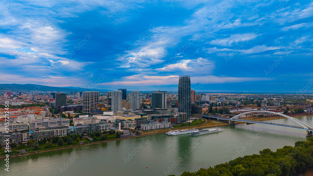 Fototapeta premium City skyline with clouds and river. View of a city skyline with tall buildings, a river, and clouds during the day. Boats are present on the water.
