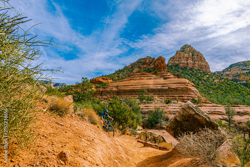 Konstfotografi A mountain biker descends a red dirt trail beneath towering sandstone buttes in Sedona, Arizona