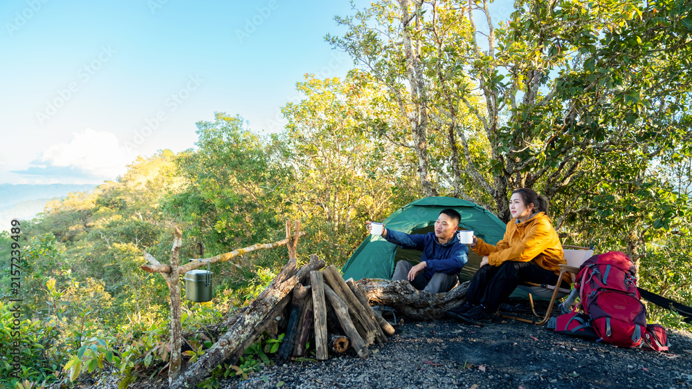 Fototapeta premium Happy Asian couple drinking hot coffee while travel nature and camping at forest mountain. Man and woman relaxing at campsite, enjoying morning beverage with outdoors active lifestyle nature therapy.