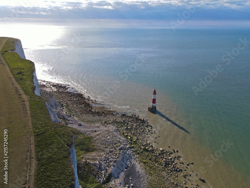 Iconic red and white striped coastal beacon Beachy Head Lighthouse at base of chalk cliffs, Eastbourne, East Sussex, England; sunny blue seaside coastal landscape on English Channel, UK