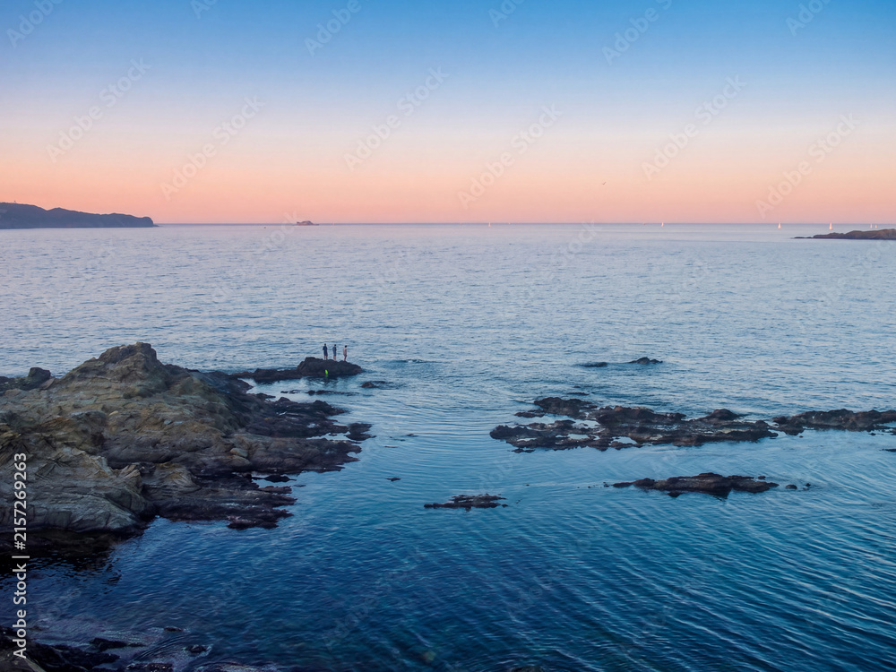 Fototapeta premium Petite crique rocheuse au crépuscule avec eau calme et transparente, paysage marin serein illuminé par des teintes roses et bleues douces