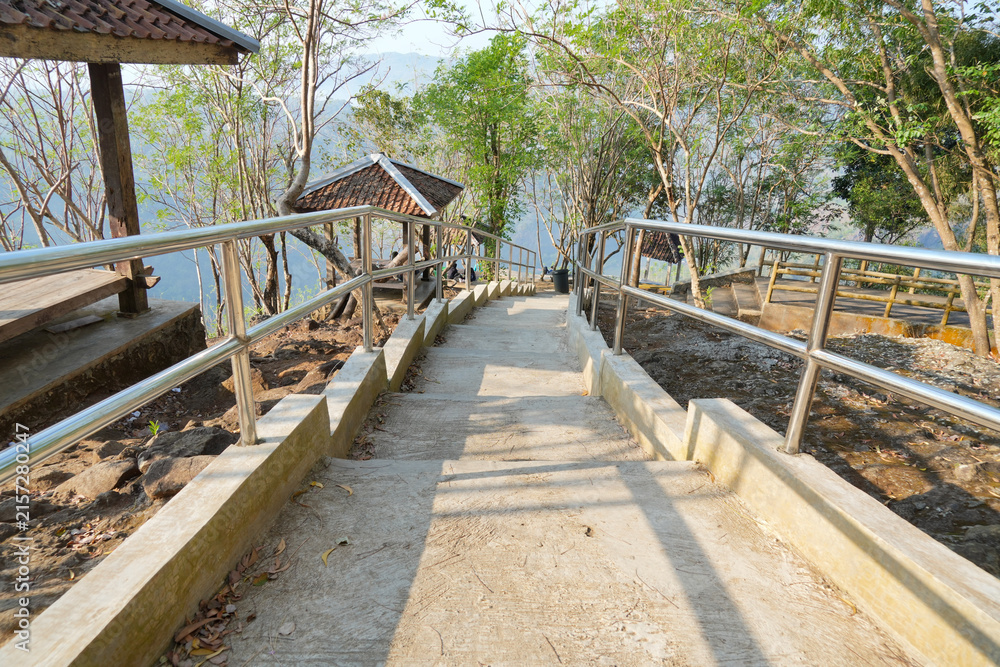 Obraz premium Concrete stairs with stainless steel handrails leading down a forested hill with wooden gazebos surrounded by lush trees in Yogyakarta, Indonesia. A scenic park trail for tourists.