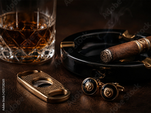 Elegant whiskey and cigar still life with crystal glass ashtray leather case and vintage cufflinks arranged on a dark wooden table with warm dramatic lighting and refined luxury lounge atmosphere