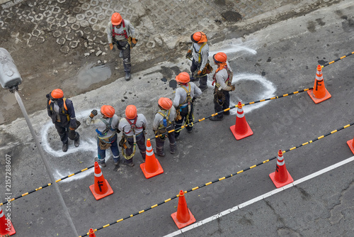 Workers gathered in an organized assembly point during an earthquake drill at a major urban building site. Wearing color-coded hard hats and full safety harnesses, the crew stands in orderly lines alo