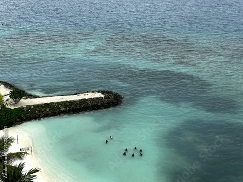 Aerial view of clear turquoise lagoon with swimmers near a rocky jetty and white sand beach