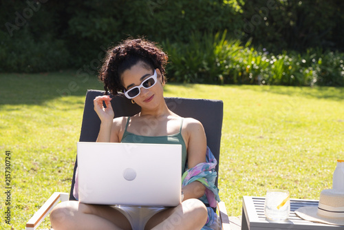 Woman sitting on cushioned lounge chair in backyard, using laptop and wearing colorful wrap
