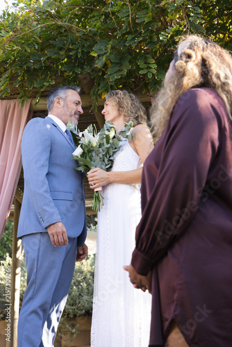 Wedding couple standing under wooden pergola in garden, bride in white lace dress holding bouquet
