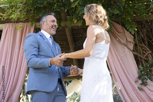 Fototapeta na wymiar Couple standing under wood arbor pink drapes garden holding hands groom blue suit bride white dress