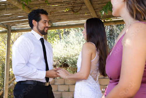 African American couple exchanging wedding rings holding hands under wooden pergola in leafy garden