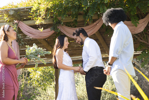 Diverse wedding couple holding hands under wood pergola with pink drape, bride wearing white gown