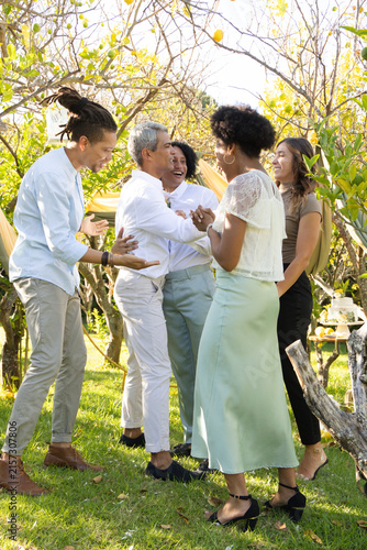 Diverse friends gathering on lawn under drape, in light attire, sharing pastries on tiered stand