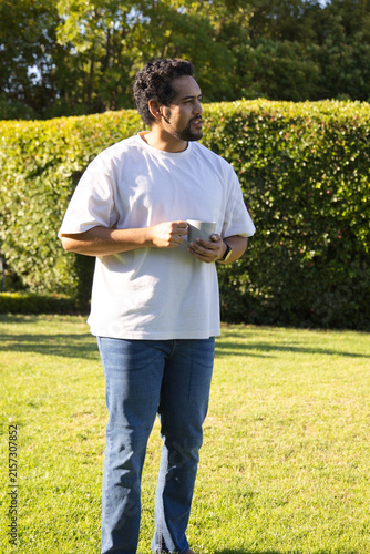 Mid adult African man standing on sunlit lawn wearing white tee holding ceramic mug looking right