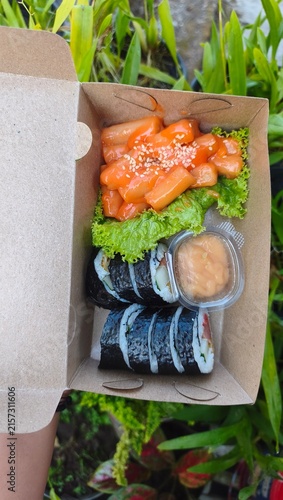 Delicious Korean street food combo featuring spicy tteokbokki rice cakes and gimbap rolls in a sustainable paper lunch box for takeout or delivery with dipping sauce and fresh lettuce