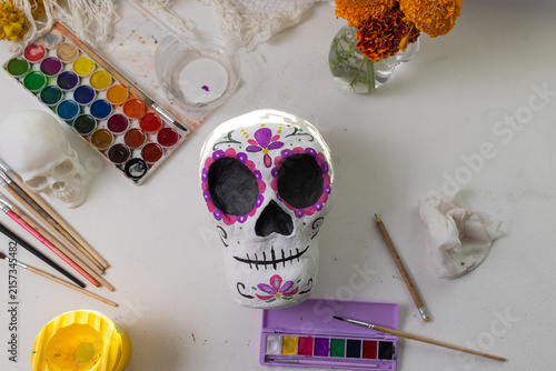 Close-up of a handmade Dia de los Muertos calavera being painted on a white studio table, showcasing the creative process from rough prep to colorful decoration with flowers and craft supplies.