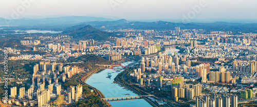Fotografie An aerial panorama of a sprawling urban city with a winding river, surrounded by distant mountain ranges under clear sky