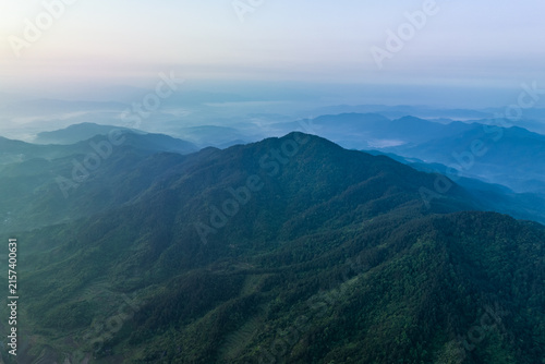 Bild auf Leinwand Aerial view of layered forested mountain ranges shrouded in soft mist at dawn, with distant hazy peaks under a pale sky