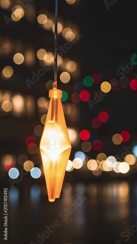 video of a glowing orange star ornament hanging outdoors at night. The star gently sways in a subtle night breeze while its inner light softly glows. In the background