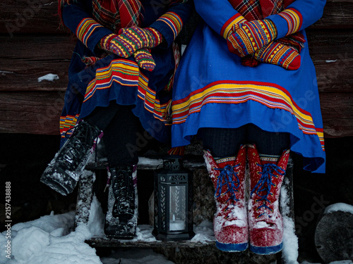 Two people wearing colorful traditional Sámi clothing, knitted mittens and winter footwear while sitting together outdoors in the snow.