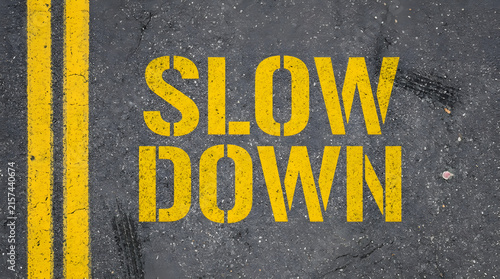 Yellow stencil text reading SLOW DOWN painted on weathered dark asphalt road surface with a double yellow line and small debris scattered around featuring pavement, marking, warning and traffic