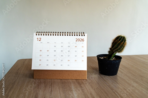 December 2026 desk calendar standing on a wooden table beside a small potted cactus. Front view office setup for time management concept.